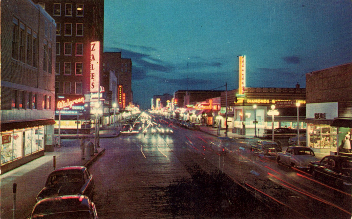 A 1950s postcard of a city street by night in America with atmospheric neon signage.