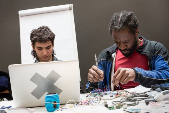 Two people hacking to create an accessible musical instrument. One sits in from of a laptop, the other uses a soldering iron. There are wires.
