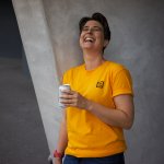 A woman with short dark hair is leaning against a grey wall. She has her head thrown back in laughter. She has a can in her hand and is wearing a yellow t-shirt..