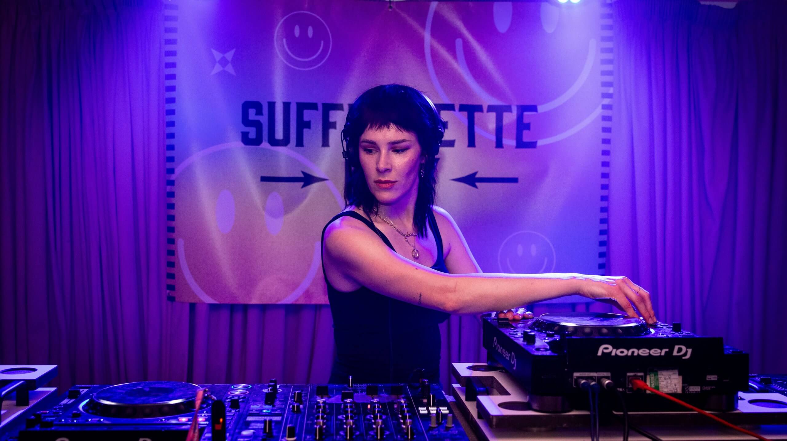 DJ Laura Jackson DJing at Suffragette City. The room is bathed in a haze of purple. She has sharply cut dark hair and high cheekbones.
