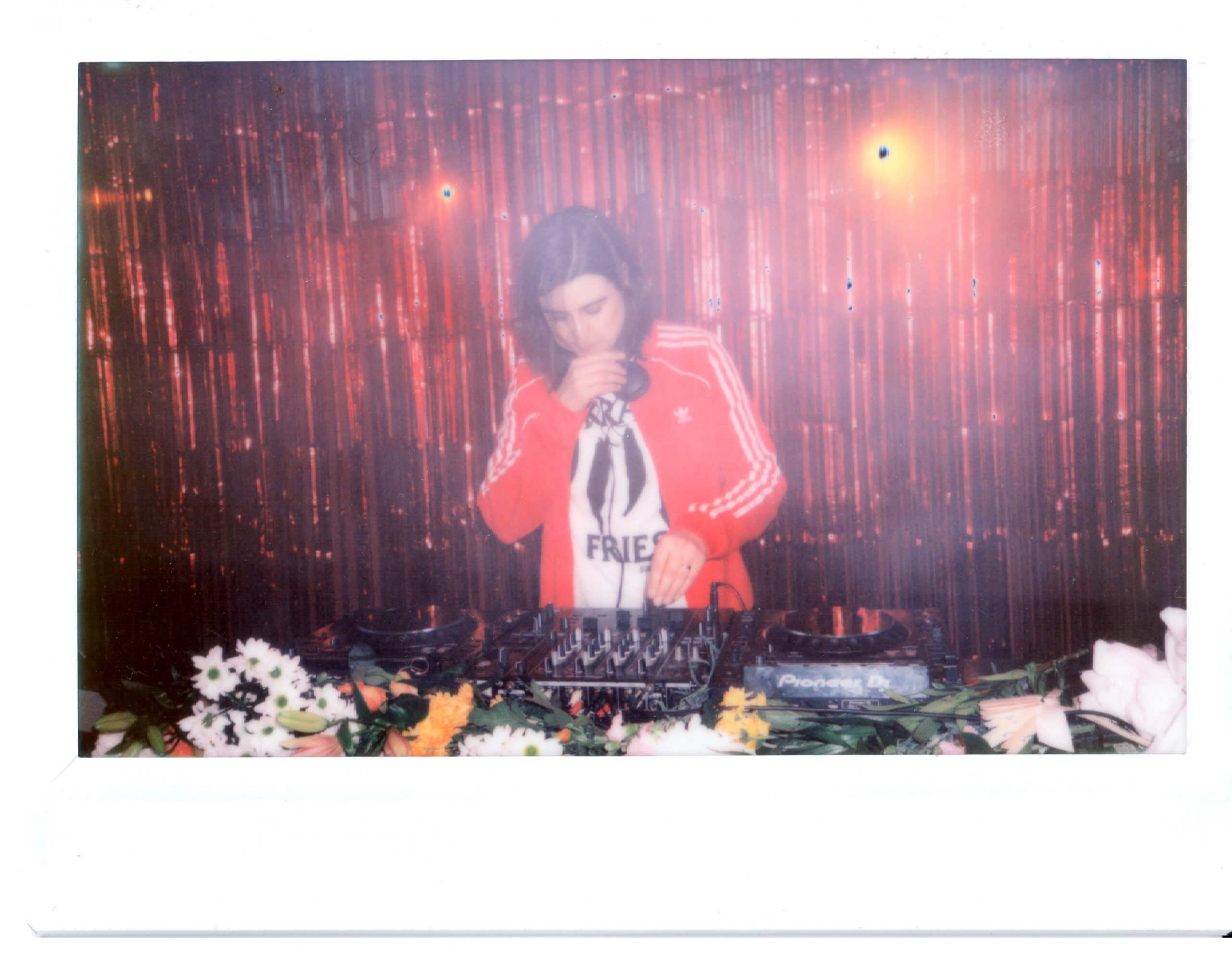 DJ with shoulder length brown hair and a red adidas top djing in front of a glittery curtain. she is using pioneer cdjs and there are flowers scattered all around the decks